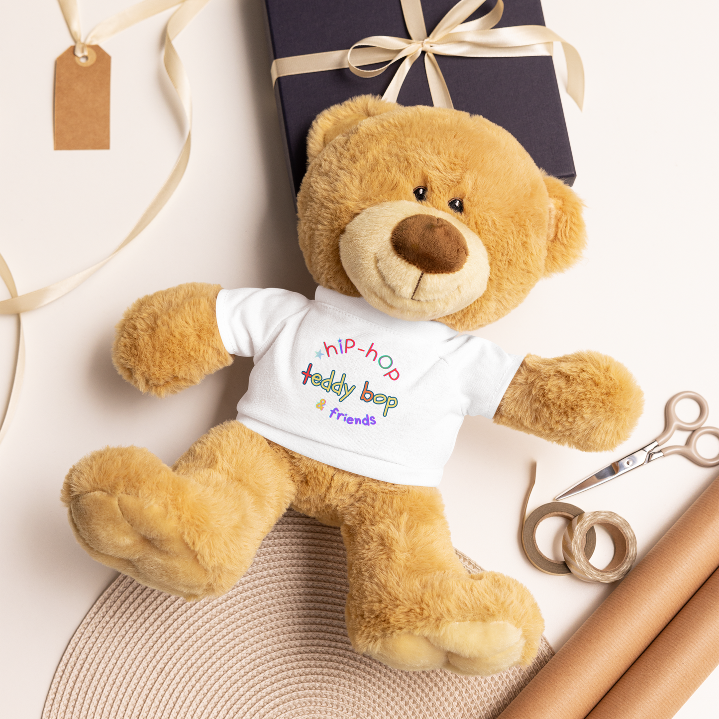 Teddy bear wearing a t-shirt with text, surrounded by gift box and scissors on a light background